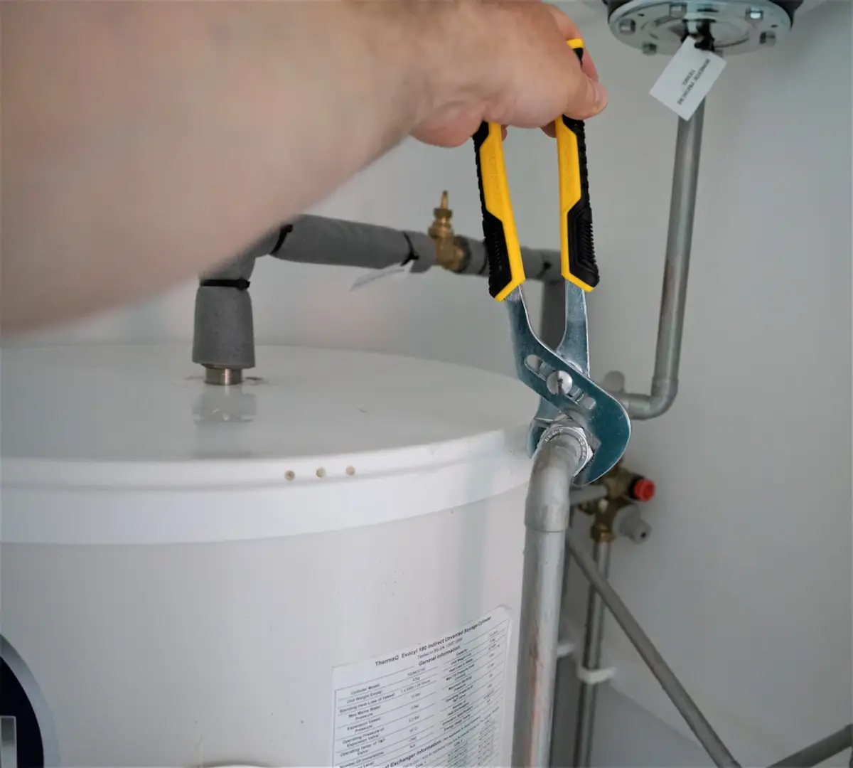 hand using pliers to tighten a pipe on a water heater tank with visible valves and plumbing connections